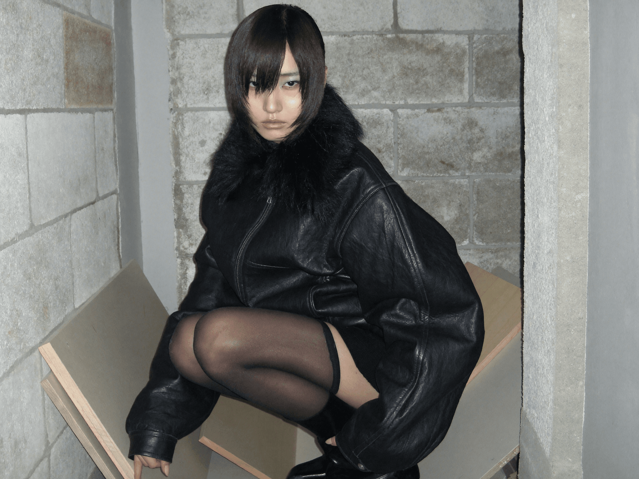 Leather jacket, black fur collar, model crouching in concrete stairwell, dark editorial, dramatic shadows