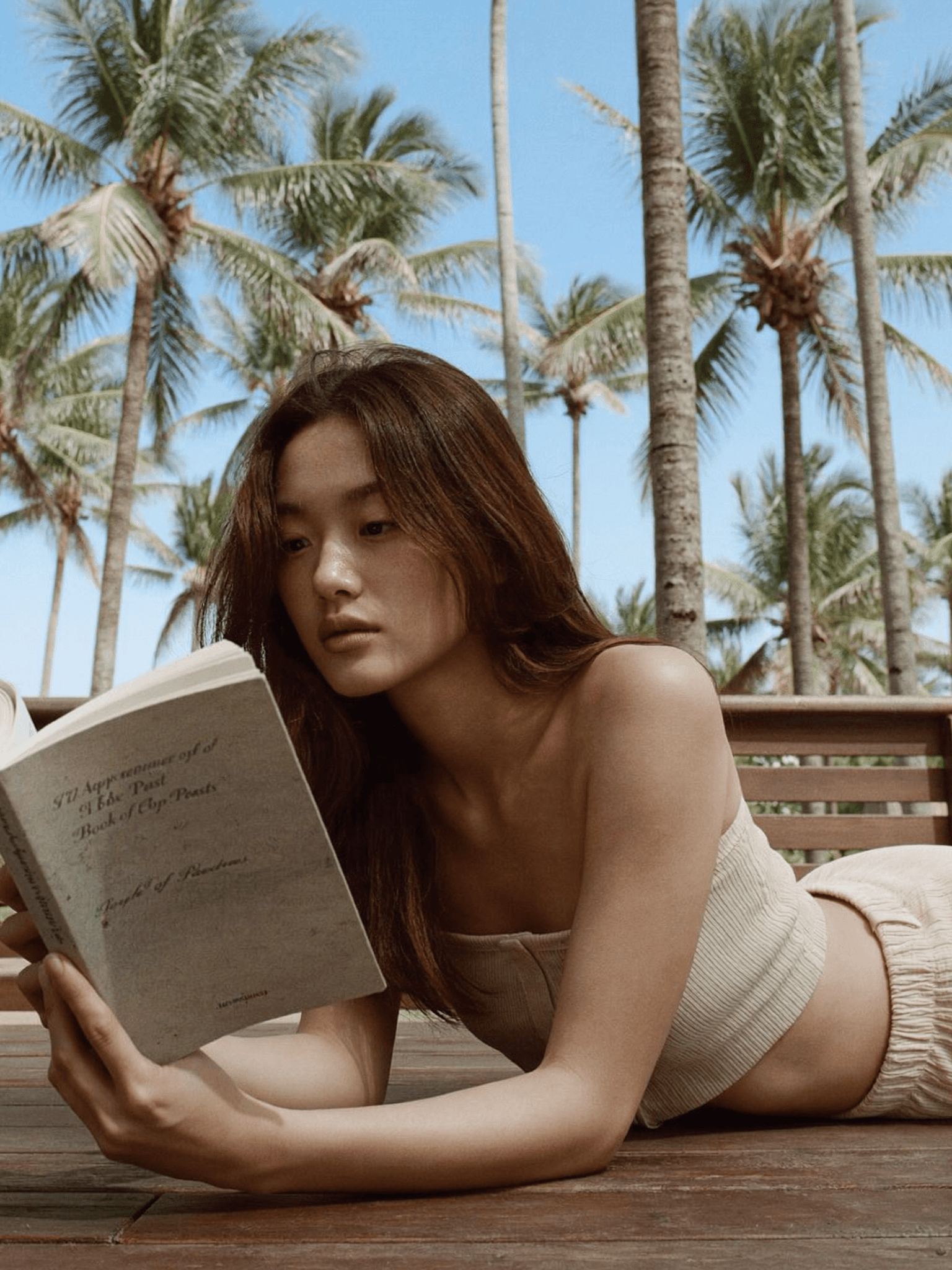 Woman reading on wooden deck by palm trees, cream bandeau, editorial lifestyle photography, Bali resort